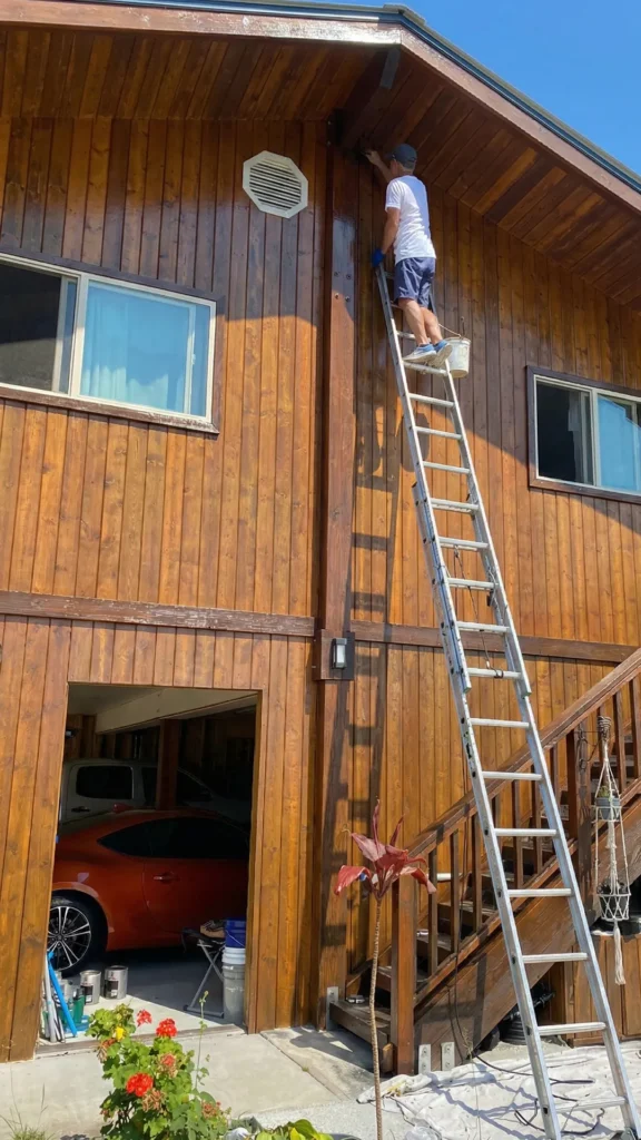 painter billy ricci on a tall ladder wood staining the side of a home